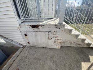 Damaged and peeling wooden storage panel under porch with rusted hardware – before repair by Flash Handy